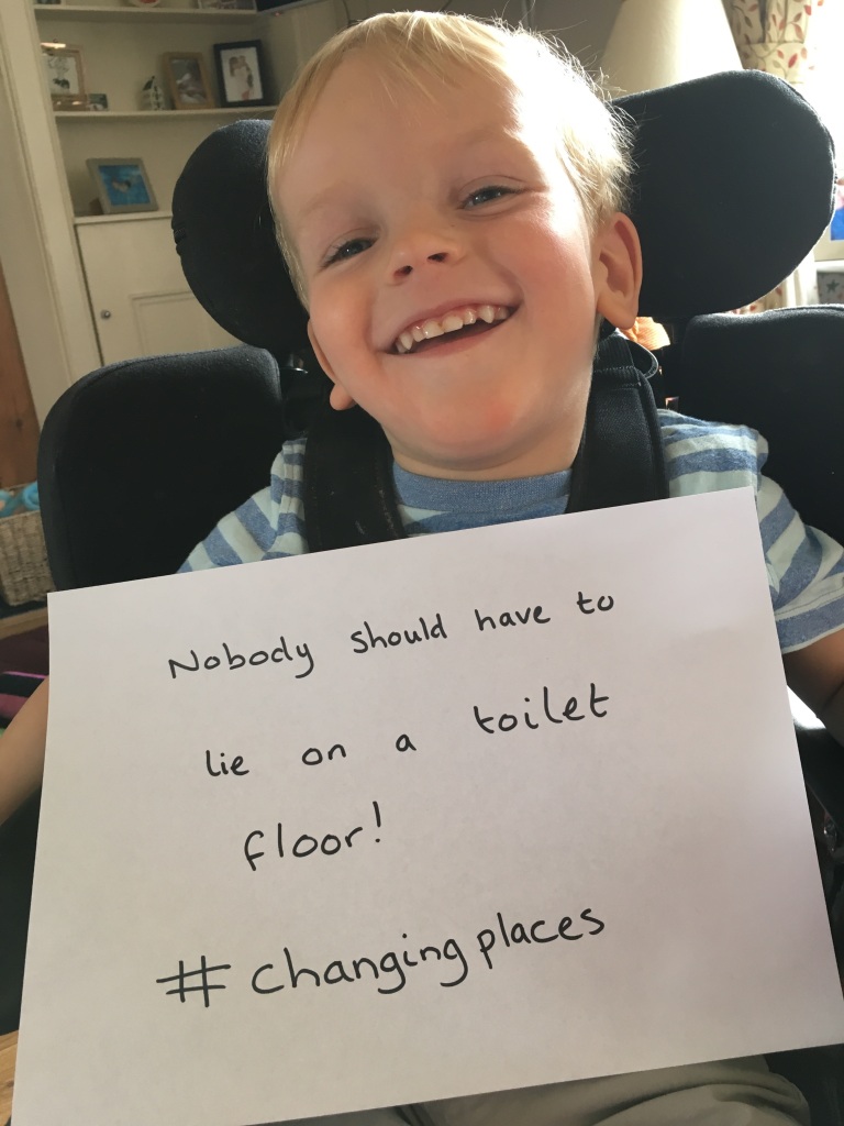 3 year old Quinns sits in his working chair smiling with a handwritten sign in front of him which reads 'Nobody should have to lie on a toilet floor! #ChangingPlaces