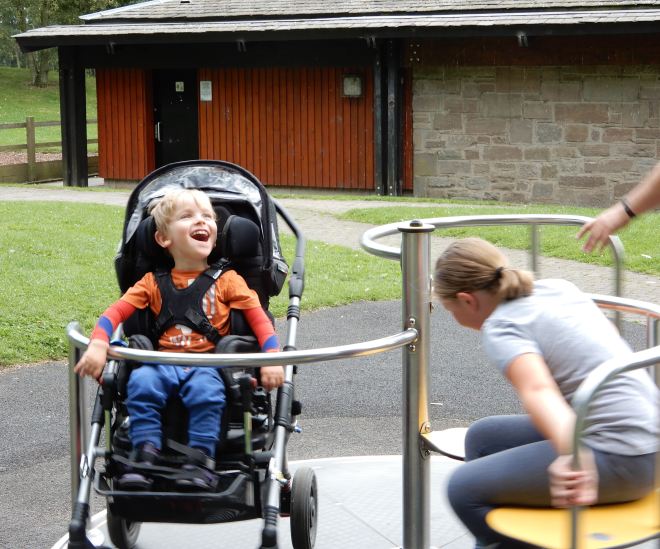 Photo of Quinns and his Big Sister enjoying an accessible roundabout with Dad pushing and a Changing Place toilet in the background.