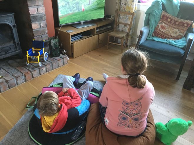 Photo of Quinns sitting in his Squiggles pack watching tv beside his Big Sister who is sitting on a bean bag. They are facing away from the camera and both are wearing hoodies.