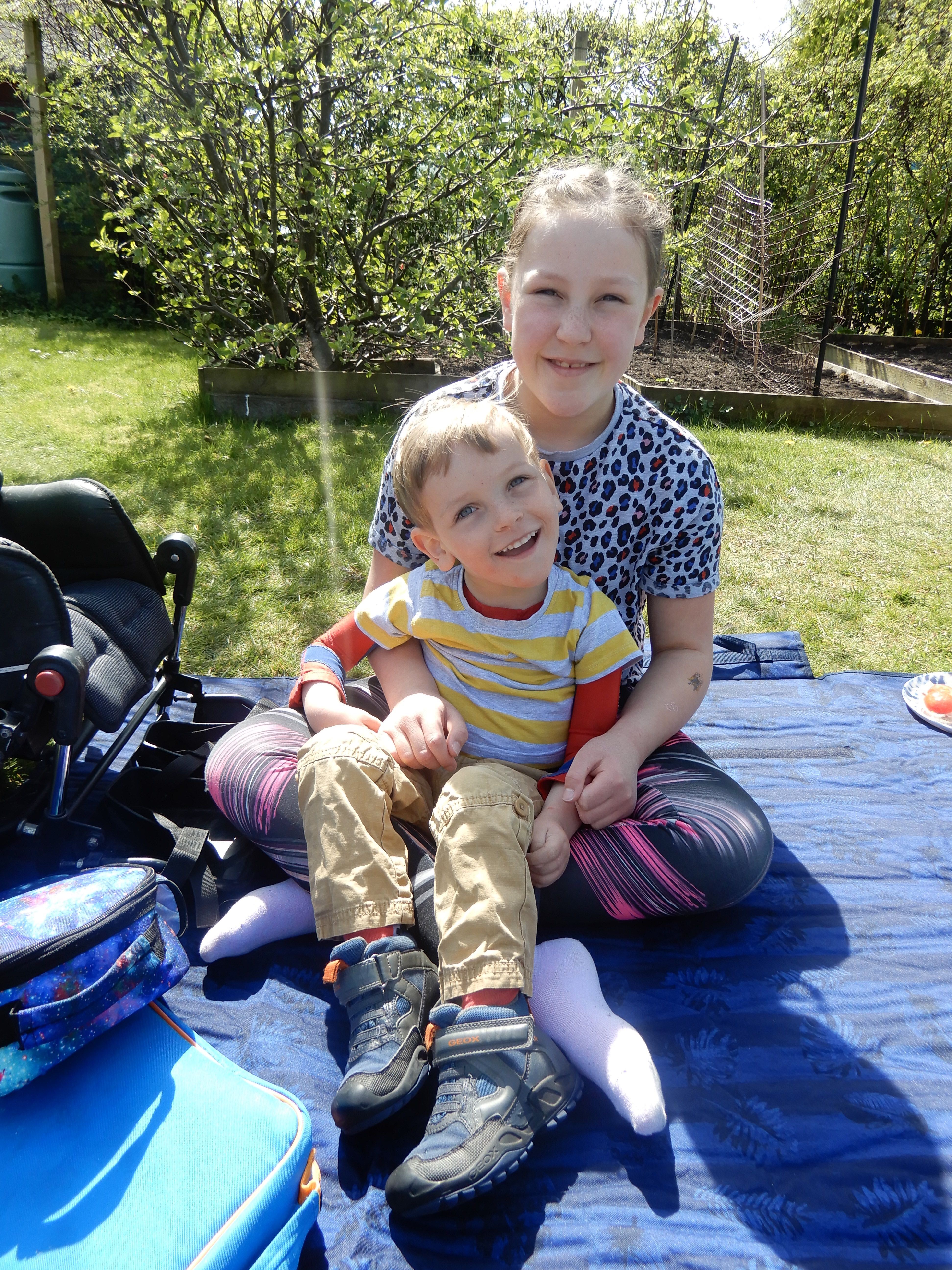 Photo of Quinns sitting on Big Sister's knee on a picnic mat with apple tree in the background.