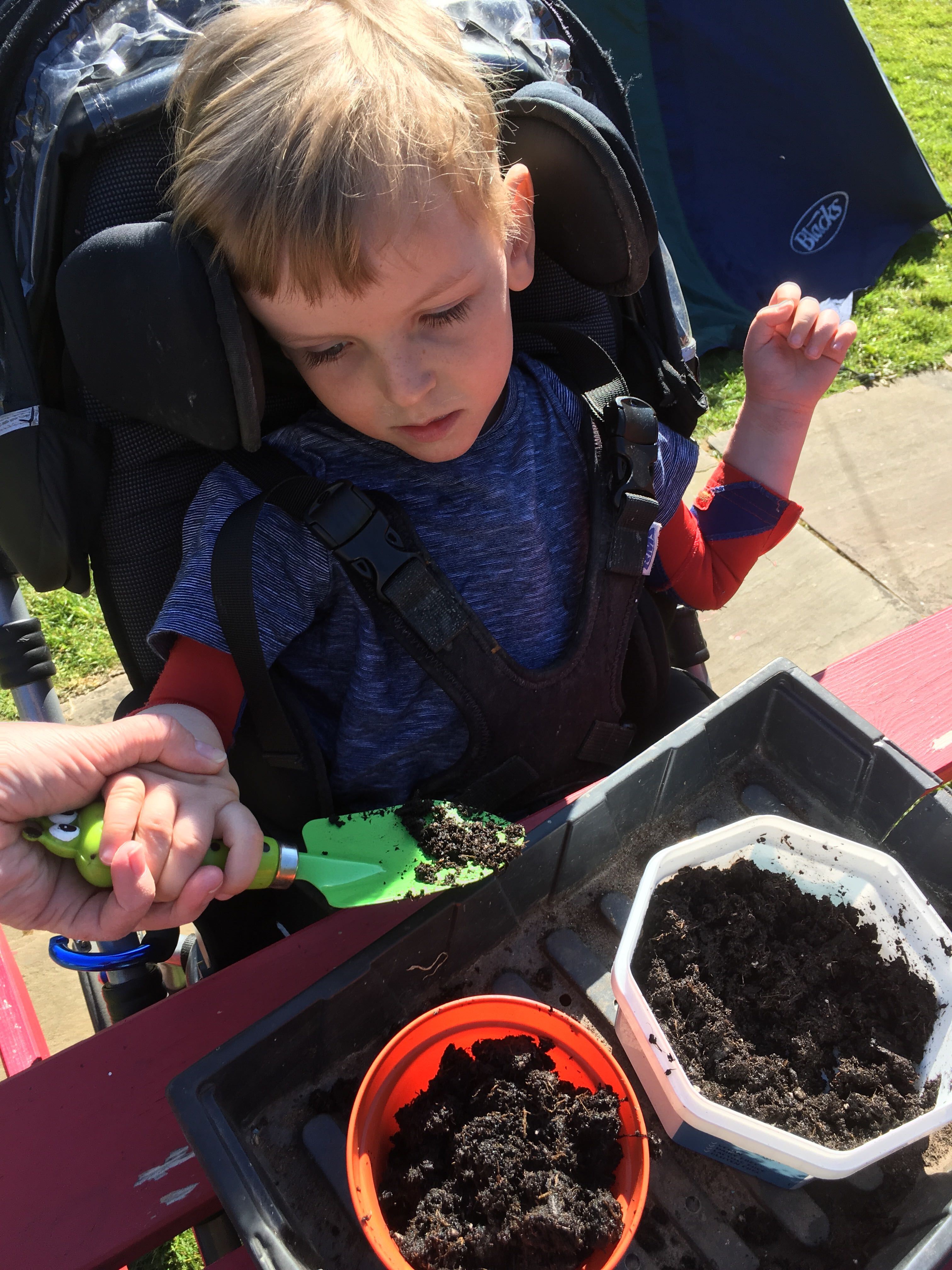 Photo of Quinns holding a green trowel moving soil from a plastic container into a plant pot guided by an adult hand