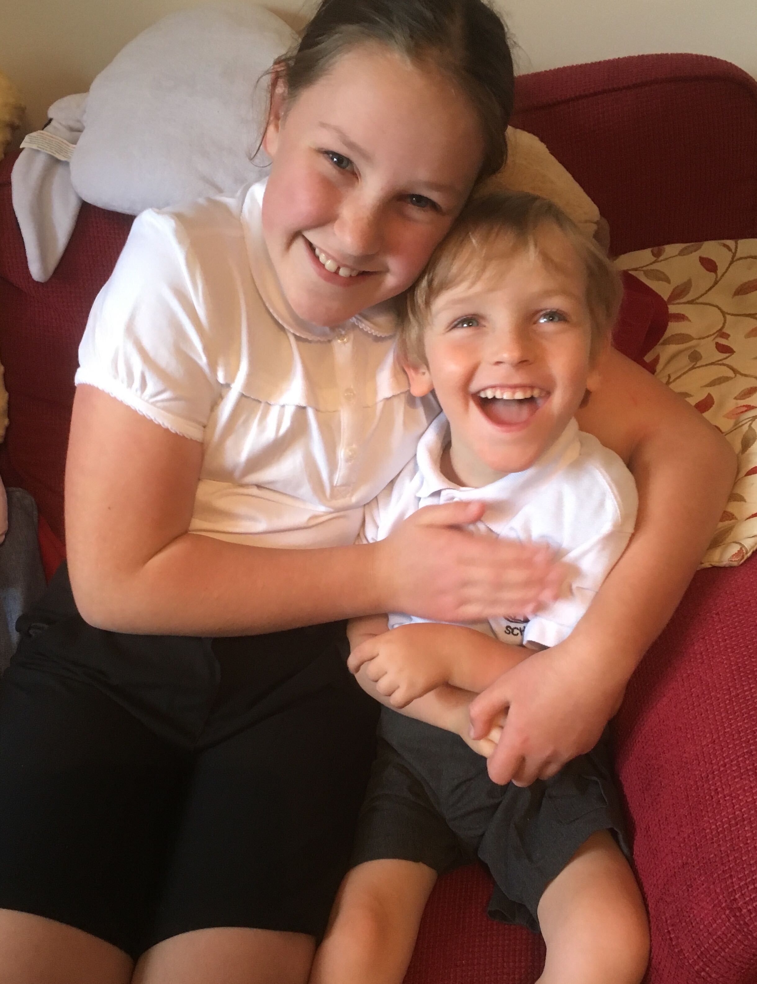Photo of Big Sister and Quinns sitting together on a red sofa both wearing dark school shorts and white polo school polo shirts. They are both smiling.