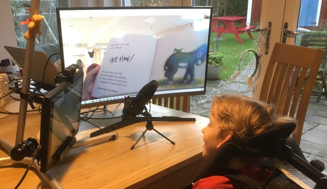 On the left sits an eye gaze computer and computer monitor sit on a table. Quinns, a 5 year old boy, sits in his chair on the right facing the screens.