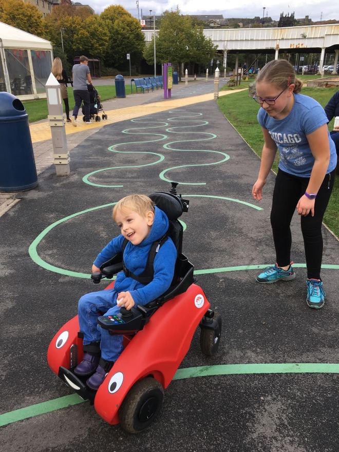Four year old Quinns drives his 'Wizzybug' small electric wheelchair with his nine year old big sister chasing him from behind.
