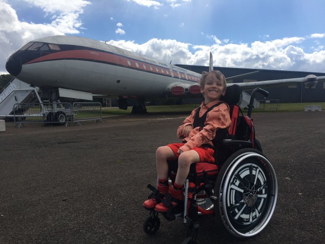 Quinns smiles his biggest smile with a large aeroplane behind him on tarmac.