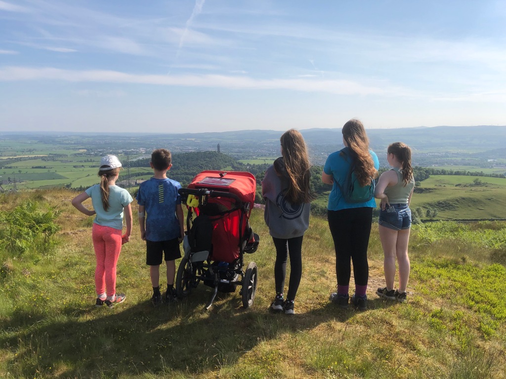 Six children with their backs to the camera including Quinns in his Delta buggy look out over Stirling. In the distance you can see the Wallace Monument. It is very small.