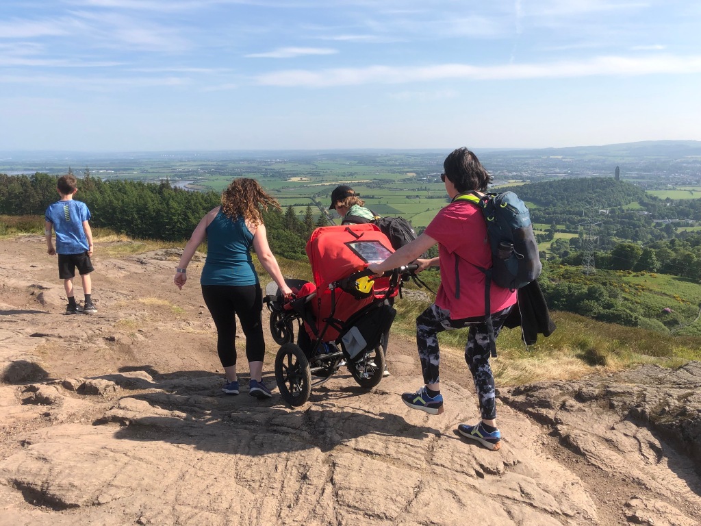 Three adult women manoeuvre Quinns in his Delta buggy across rocky terrain with a view of the Wallace Monument in the distance. A seven year old boy is in front of the Delta. 