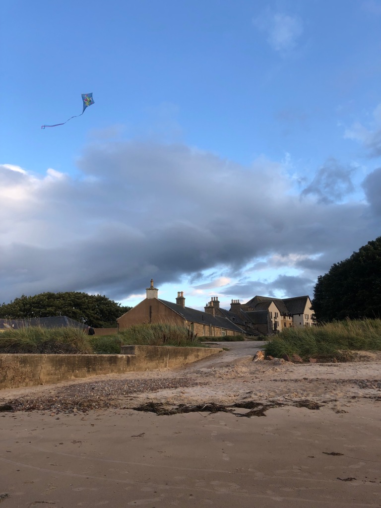 A kite flies high above the clouds and some cottages by the beach.