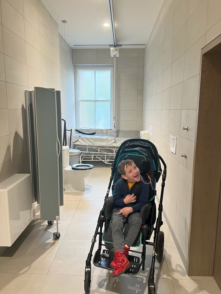 Quinns smiles in the centre of a narrow Changing Places facility with ceiling track hoist, adjustable height changing bed, toilet and screen.