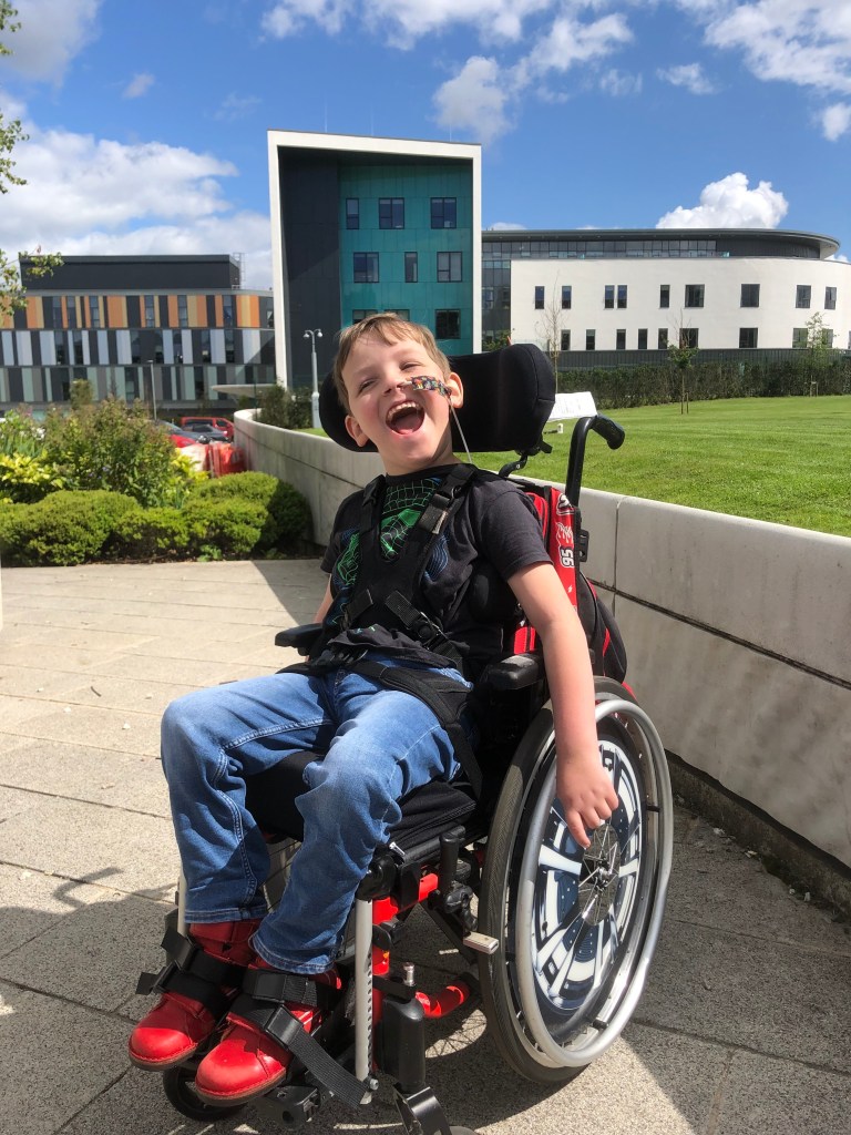 Quinns smiles sitting in his wheelchair. He has on red shoes, jeans and a black t-shirt. The RHCYP Edinburgh is in the background.