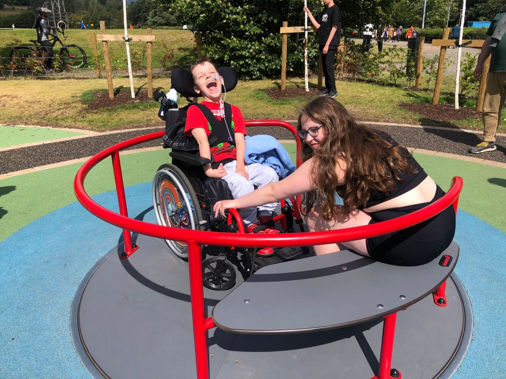 Quinns throws his head back in excitement as Anita holds on tight while the two of them spin on a wheelchair accessible roundabout.