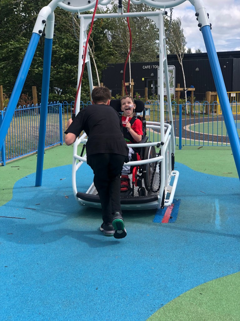 Quinns grins a broad grin as his friend pushes him on the wheelchair accessible swing.