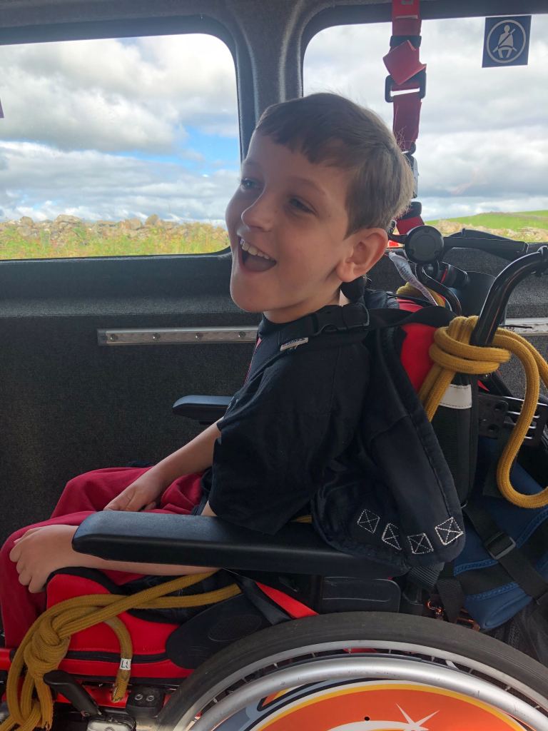 Quinns smiles in his wheelchair which is strapped into the Bendrigg bus on the way to caving. Yellow ropes can be seen attached to the side of his chair. 