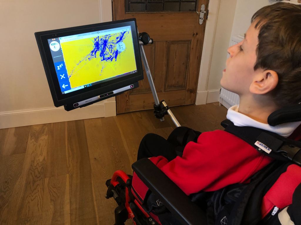 The photo is taken over Quinns shoulder. He is sitting in his wheelchair looking at the screen of his eye gaze which shows a bright yellow and blue artwork.