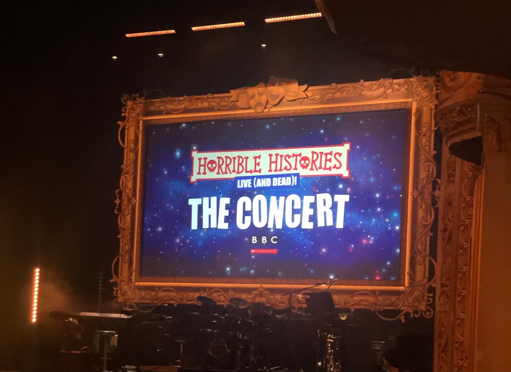 Close up of the screen on the stage at the Theatre Royal in Glasgow. It reads Horrible Histories, Live (and Dead)! The Concert BBC.