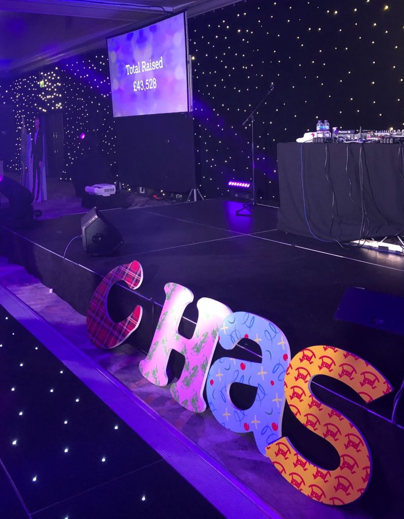 Screen reading total raised £43,528 in the background. Giant patterned wooden letters spelling CHAS in the foreground.