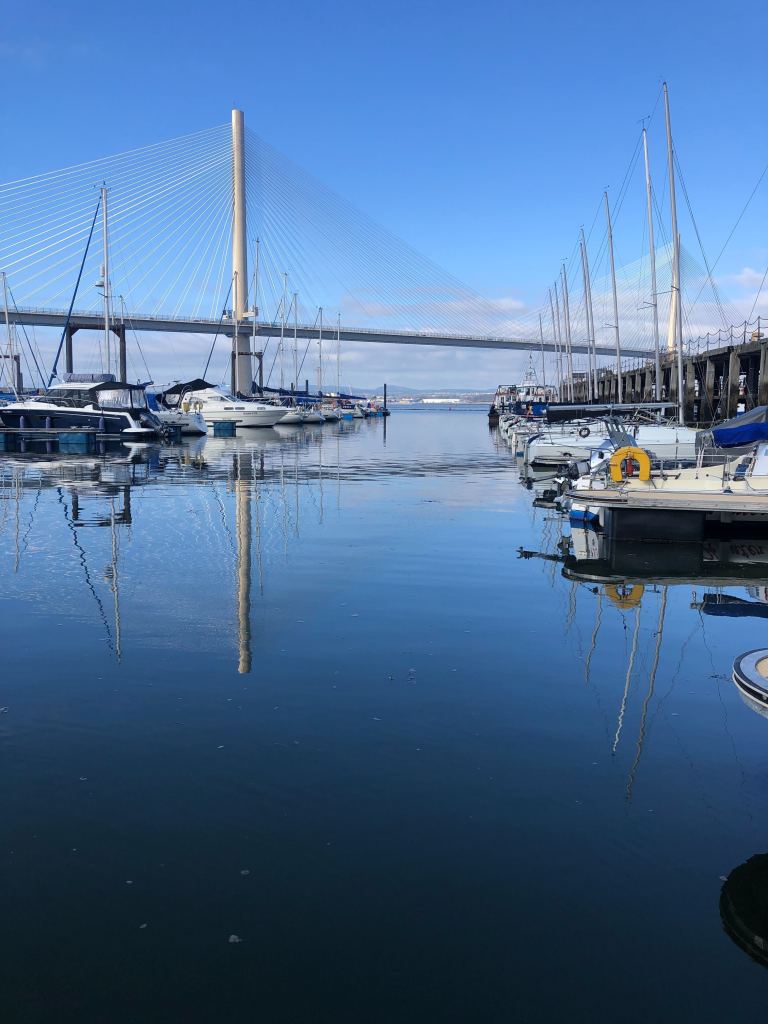 Boats in the harbour with the new Queensferry Crossing in the background. The sky and the water are the same blue and the reflections of boats and bridge are really clear.