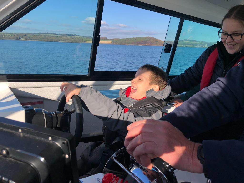 Quinns is smiling as he holds the steering wheel for the Wet Wheels boat. Big Sister is smiling and watching him from behind.