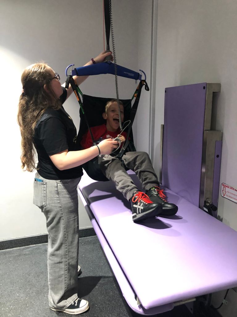 Anita is hoisting Quinns in his sling onto a purple adjustable height changing bench.