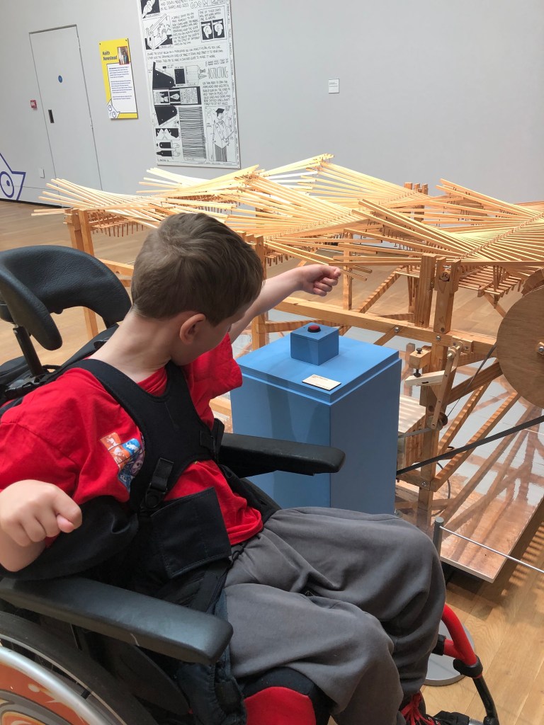 Quinns is reaching to push a red button to make interconnecting wooden slats move.